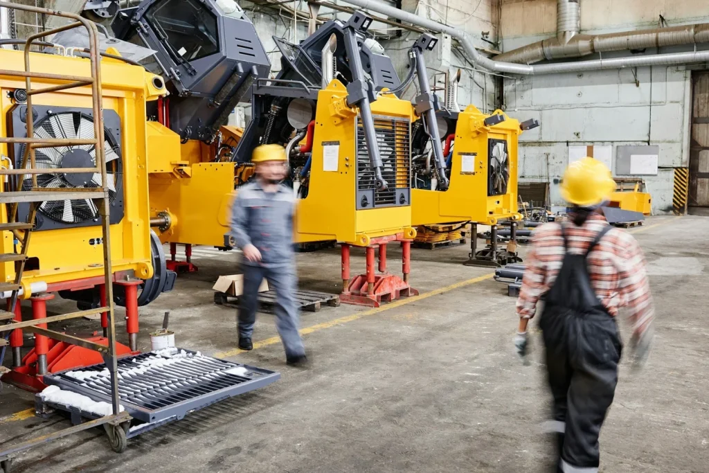Workers wearing hard hats beside large yellow machinery on a factory floor.