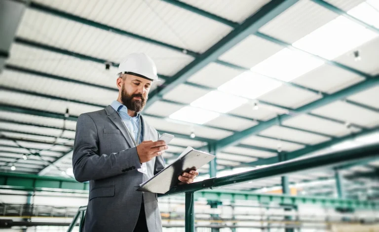 A man wearing a hard hat reviews a clipboard inside an industrial building.